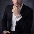 A man in a formal black suit and white shirt showcasing a silver diamond-encrusted watch on his wrist.